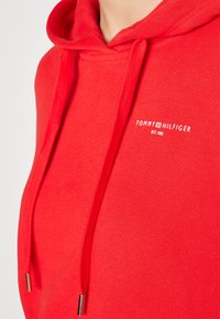 Sweat à capuche rouge avec cordons, avec petit logo blanc Tommy Hilfiger et texte "EST. 1985" sur la poitrine.