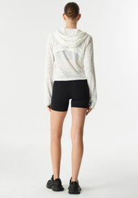 Haut à capuche léger et à manches longues de couleur blanche avec un design texturé et un panneau en maille à l'avant, associé à un short noir et des chaussures de sport noires.