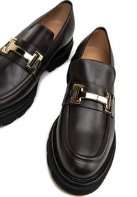 Hnědé kožené mokasíny s detaily zlatého tónu na sponě. Mají robustní černou gumovou podrážku a hladký povrch. Minimalistický design.