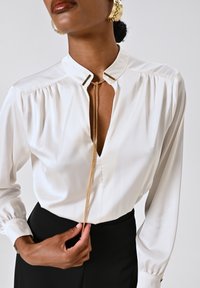 Blusa bianca in satin con scollatura a V profonda e dettaglio in catena dorata. Caratterizzata da maniche lunghe con polsini arricciati e dettagli eleganti al colletto.