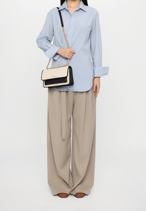 Femme portant une chemise à rayures bleu clair et un pantalon beige à jambes larges, tenant un sac à bandoulière crème et noir avec une chaîne dorée.