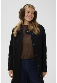 Čierny kabát s textúrou a veľkými �čiernymi gombíkmi, doplnený hnedým svetrom. Modelka má na ušiach hnedé kožušinové nausnice. Viditeľné modré džínsové nohavice.