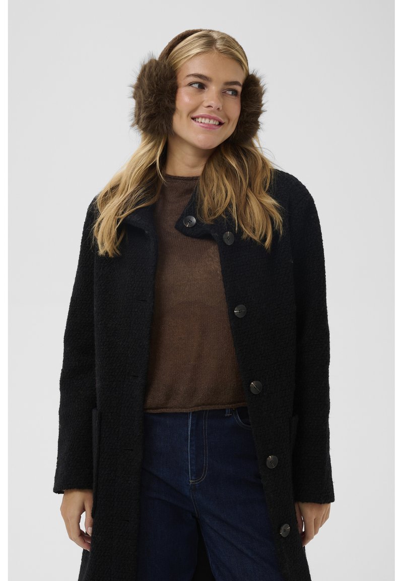 Čierny kabát s textúrou a veľkými čiernymi gombíkmi, doplnený hnedým svetrom. Modelka má na ušiach hnedé kožušinové nausnice. Viditeľné modré džínsové nohavice.