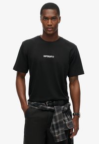 Sort bomulds t-shirt med et hvidt "SUPERDRY®" logo, parret med sorte bukser og en gråternet skjorte bundet om taljen.