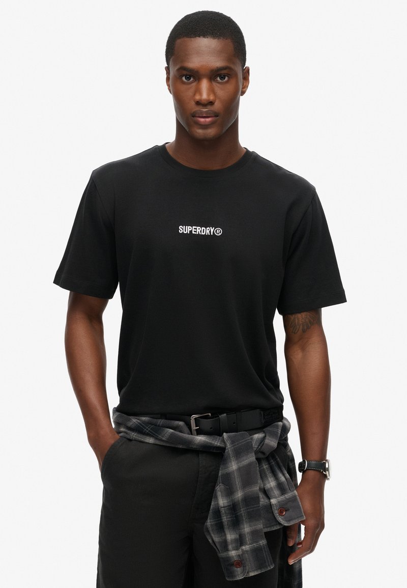 Sort bomulds t-shirt med et hvidt "SUPERDRY®" logo, parret med sorte bukser og en gråternet skjorte bundet om taljen.
