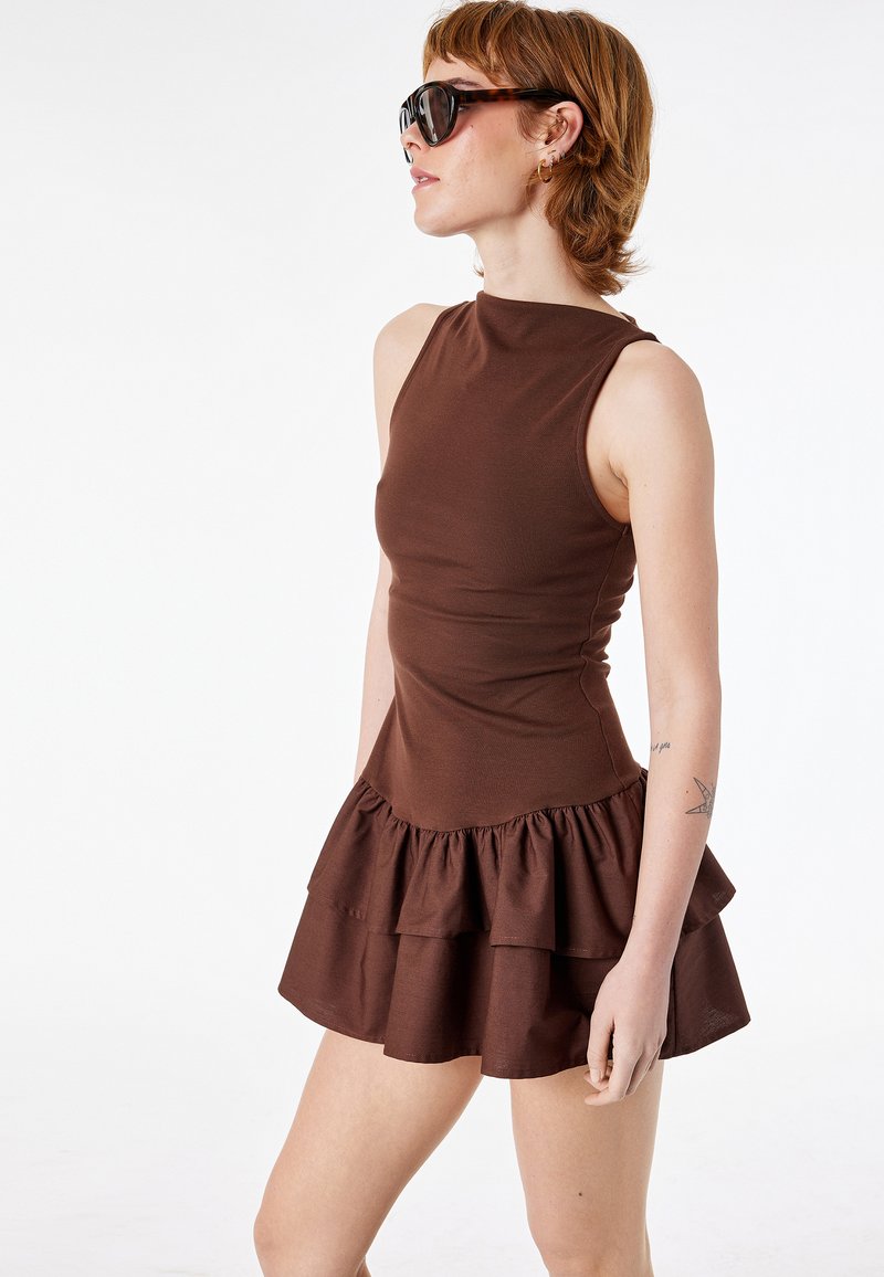 Junge Frau mit einem ärmellosen braunen Kleid mit einem Rock aus Rüschen, großen schwarzen Sonnenbrillen und kurzen hellbraunen Haaren, seitlich gewandt.