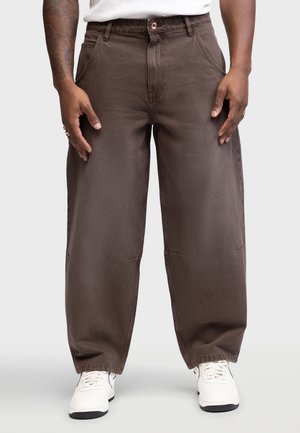 Hombre con pantalones marrones sueltos y zapatillas blancas, de pie con las manos relajadas a los lados sobre un fondo liso.