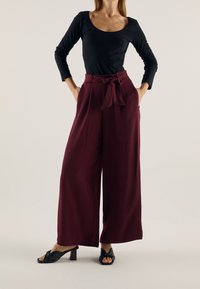 Pantalon large bordeaux avec taille à nouer, tissu texturé, poches latérales, associé à un haut noir à manches longues et des sandales à talons noires.