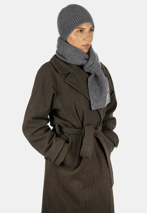 Frau trägt einen dunkelbraunen, mit Gürtel ausgestatteten Mantel, graue Strickmütze und passenden grauen Strickschal, schaut zur Seite und hat die Hände in den Taschen.
