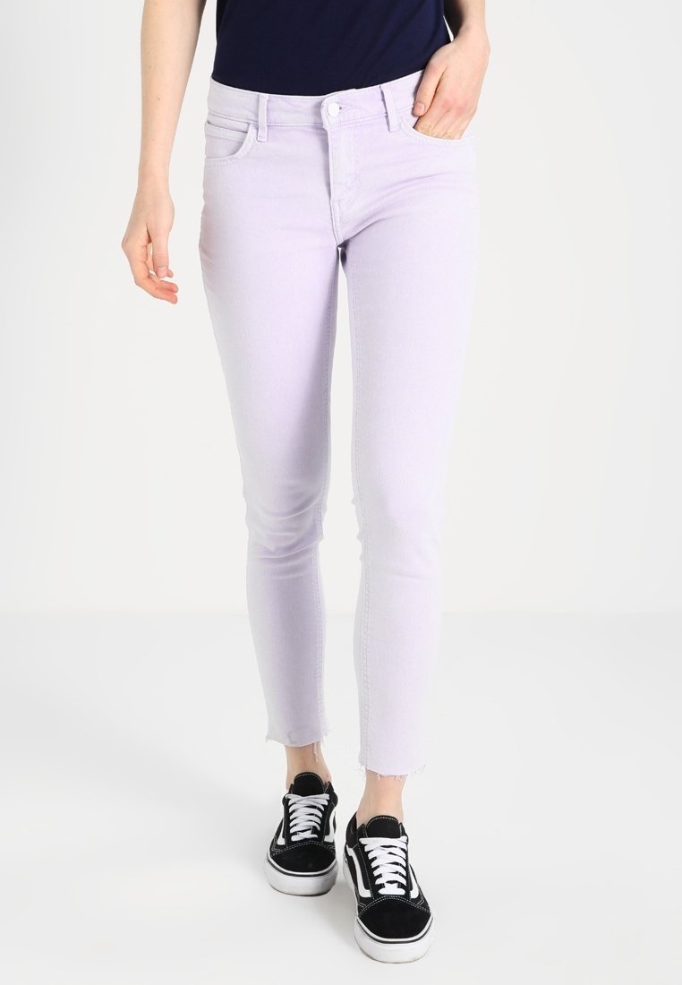 Pantalones vaqueros ajustados de color púrpura claro, confeccionados en denim, con un diseño clásico de cinco bolsillos y dobladillos deshilachados, combinados con zapatillas negras.