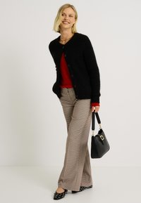 Cardigan noir en tricot avec des boutons, pull rouge en dessous, pantalon à carreaux et sac à main noir. Chaussures noires avec des pois blancs.