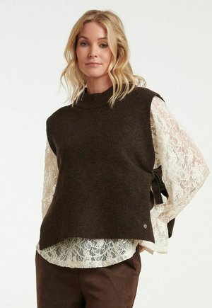 Blonde vrouw draagt een donkerbruine mouwloze gebreide vest over een witte kanten blouse met lange mouwen, staand tegen een effen achtergrond.
