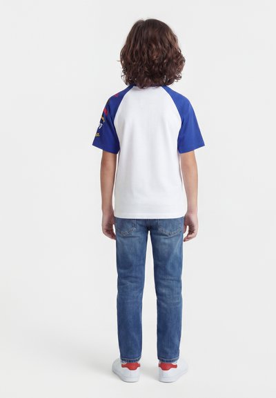 Παιδί με μαλλιά μέχρι τον ώμο και σγουρά, στέκεται γυρισμένο σε άλλον, φοράει λευκό και μπλε κοντομάνικο πουκάμισο, μπλε τζιν και λευκά αθλητικά παπούτσια με κόκκινες λεπτομέρειες.