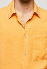 Uomo che indossa una camicia color arancione brillante con il colletto sbottonato e una tasca sul petto, mostrando un po' di peli sul petto e nei nei sul collo.
