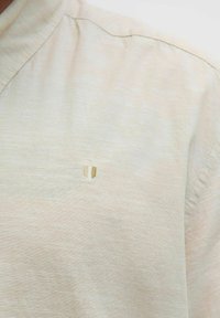 Chemise beige à manches courtes avec une finition texturée ; présente un petit emblème en forme de bouclier subtil en or clair sur la partie gauche de la poitrine.