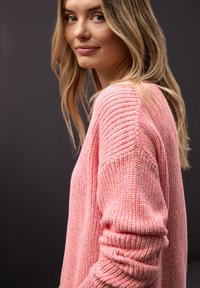 Jeune femme aux longs cheveux blonds portant un pull en tricot rose, regardant légèrement sur le côté sur un fond gris foncé.