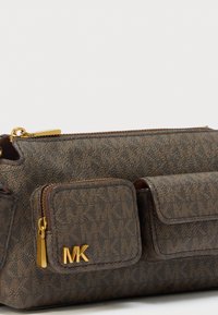Smeđa torba od teksturirane tkanine sa zlatnim detaljima, s kopčanjem na patentni zatvarač i dva prednja džepa. Istaknuti logo "MK".