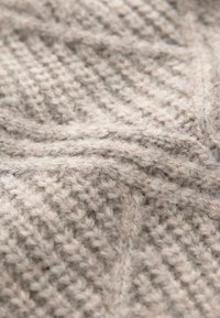 Grobmaschiges Strickgewebe in hellbeige mit einem strukturierten Fischgrätenmuster. Weiche, flauschige Textur mit sichtbaren Fasern und genähten Details.
