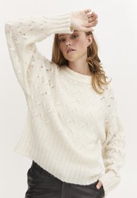 Cremfarbener Strickpullover mit gerippten Bündchen und Saum, der ein offenes Sternmuster aufweist. Weiche Textur, lockere Passform und runder Ausschnitt.
