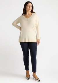 Cremefarbener V-Ausschnitt-Pullover mit langen Ärmeln, geripptem Saum und Knopfakzenten an den Bündchen, kombiniert mit dunkelblauen Slim-Fit-Hosen und silbernen Absatzschuhen.