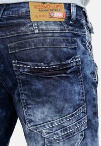 Jeans in denim con un lavaggio blu sbiadito, dotati di un'etichetta in pelle con la scritta "KOSMO•LUPO" e numerose tasche cucite con filo a contrasto.