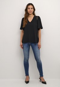 Top nero a V con maniche corte a svaso e dettagli decorativi sulle spalle, abbinato a jeans aderenti blu e décolleté nere a punta.