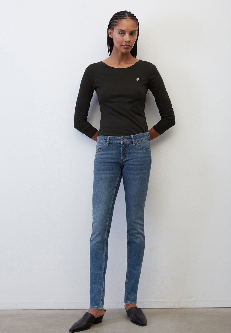 Czarna koszula z długim rękawem, dopasowany fason, w zestawieniu z niebieskimi rurkami. Gładki materiał z subtelnym logo na koszuli. Czarne buty wsuwane.