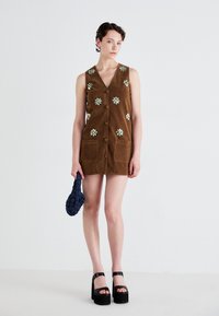 Brown suede sleeveless dress with flower embellishments, button front, and two side pockets. Paired with thick black platform shoes and a braided bag.