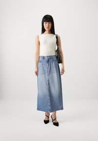 Un débardeur blanc à côtes associé à une jupe maxi en denim bleu clair, avec une coupe droite et un léger dégradé. Des talons aiguilles noirs à lanières et un sac à main.