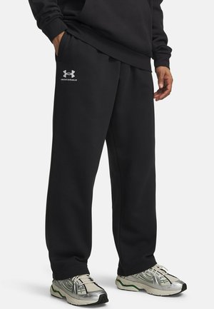 Person trägt schwarze Under Armour Jogginghose mit den Händen in den Taschen und silberne Sportschuhe mit grünen Akzenten.