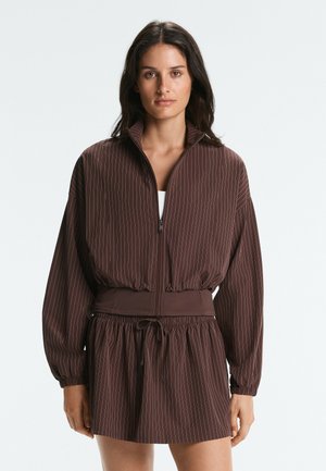 Frau trägt eine braune Nadelstreifen-Zipjacke mit elastischem Bund und passenden Kordelzugrock, steht vor einem schlichten Hintergrund.