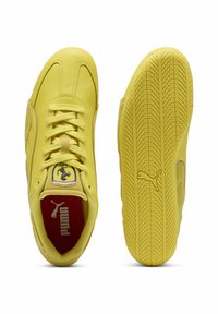 Zapatillas de cuero amarillas con una superficie texturizada, que cuentan con un logo de Ferrari en la lengua, plantilla roja y suela de goma con un patrón de tracción.