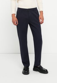 Sötétkék vászon nadrág, egyenes szabással, gombos zárással és oldalsó zsebekkel. Fekete slip-on cipőkkel viselve.