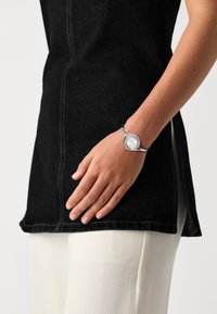 Schwarzes Denim-Tunika mit Seitenschlitz, kombiniert mit einer silbernen Metalluhr mit weißem Ziffernblatt und einem flexiblen, dekorativen Armband.