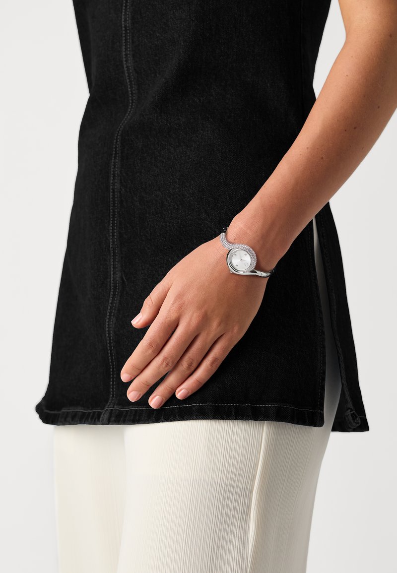 Schwarzes Denim-Tunika mit Seitenschlitz, kombiniert mit einer silbernen Metalluhr mit weißem Ziffernblatt und einem flexiblen, dekorativen Armband.