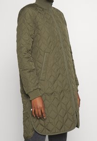 Chaqueta larga acolchada de color verde oliva con un alto cuello, bolsillos laterales y puños acanalados. Presenta un patrón de diamantes en la tela. Cierre de cremallera.