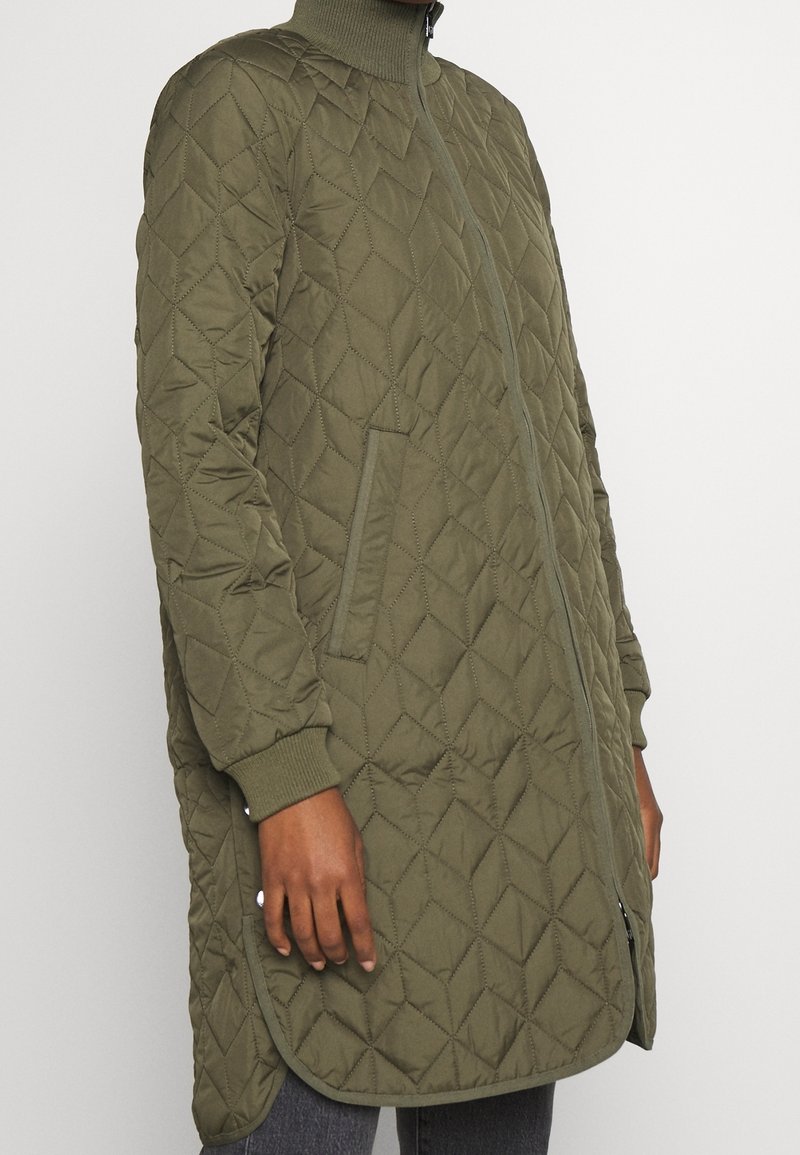 Chaqueta larga acolchada de color verde oliva con un alto cuello, bolsillos laterales y puños acanalados. Presenta un patrón de diamantes en la tela. Cierre de cremallera.