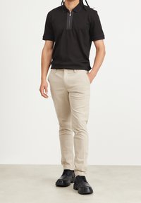 Černé polokošile s límcem na zip, doplněné světle béžovými kalhotami a černými šněrovacími botami, na neutrálním pozadí.