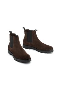 Un par de botas Chelsea de ante marrón oscuro con paneles laterales elásticos negros y suelas de goma negra tipo lug, mostradas sobre un fondo blanco.
