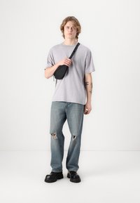 Grå oversized t-skjorte, lyseblå ripped jeans, svart midjeveske, kraftige svarte plattformsko, enkel bakgrunn.