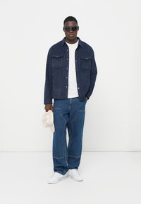 Jeansjacke mit vertikalen Streifen, weißes T-Shirt, lässige blaue Jeans, weiße Sneakers und eine cremefarbene Mütze in der Hand; Sonnenbrille vervollständigt das Outfit.