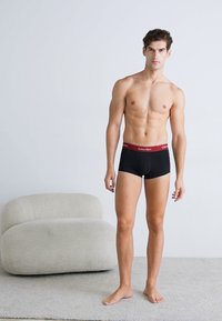 Zwarte boxershorts met een rode tailleband met daarop de merknaam, gedragen door een model, op een lichtgekleurde achtergrond met zachte meubels.