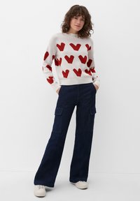 Maglione lavorato a maglia con motivi a cuore rossi su sfondo crema; abbinato a pantaloni cargo larghi blu scuro; modello in sneakers bianche.