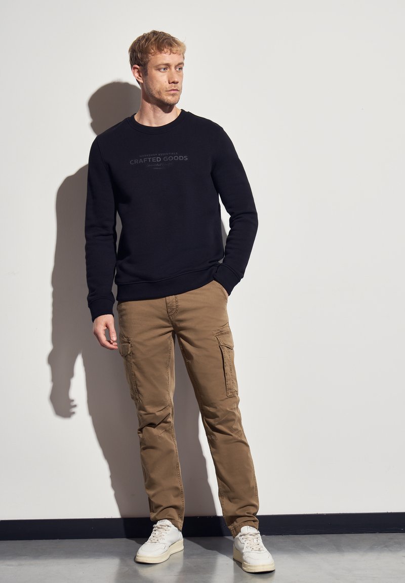 Man staat tegen een witte muur, draagt een zwarte trui met de tekst "Crafted Goods", beige cargobroek en witte sneakers, kijkt opzij.