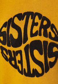 Sudadera amarilla con texto negro en relieve que dice "SISTERS" en un estilo divertido y invertido. Textura de tela suave; diseño informal.