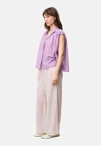 Lila Kurzarmbluse mit lockerer Passform und Knopfdetail, kombiniert mit gestreiften, weit geschnittenen Hosen in Pastellfarben. Das Schuhwerk ist in Beige.