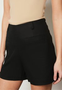 Skräddarsydda svarta shorts med en slät textur, utrustade med framfickor och rena linjer. Midjebanden är platta och skapar en slimmad silhuett.