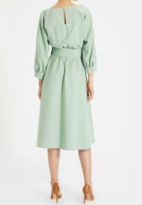 Miętowa sukienka z kluczykiem na plecach, bufiastymi rękawami, talią cinched i spódnicą o długości do łydki. Gładki materiał z subtelnym drapowaniem.