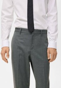 Pantalons gris sur mesure en tissu lisse, présentant une coupe slim, des poches latérales et un design à l'avant plat, associés à une chemise blanche et une cravate.