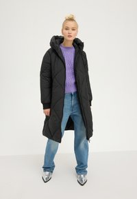 Młoda osoba stoi ubrana w długi czarny pikowany płaszcz, lawendowy sweter z grubym splotem, jasnoniebieskie dżinsy i metaliczne, szpiczaste buty na białym tle.